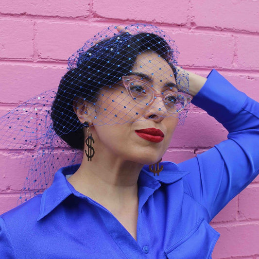 Woman in blue outfit stands in front of pink brick wall wearing "Dollar Dollar Bill" 18K Gold Statement Earrings shaped as a dollar sign featuring Black Obsidian rough cut crystals