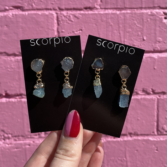Two pairs of Crystal drop Earrings. 18K Gold Statement Earrings featuring Aquamarine rough cut crystals. On black Scorpio earring backing cards held in front of a pink brick wall.