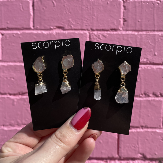 Two pairs of Crystal drop Earrings. 18K Gold Statement Earrings featuring Clear Quartz rough cut crystals. On black Scorpio earring backing cards held in front of a pink brick wall.