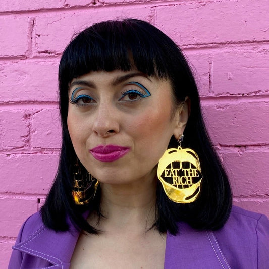 18K Gold Statement Earrings shaped as a mouth with the words "Eat The Rich" between the top and bottom row of teeth featuring Amethyst rough cut crystals. Modelled by Margeaux in front of a pink brick wall.