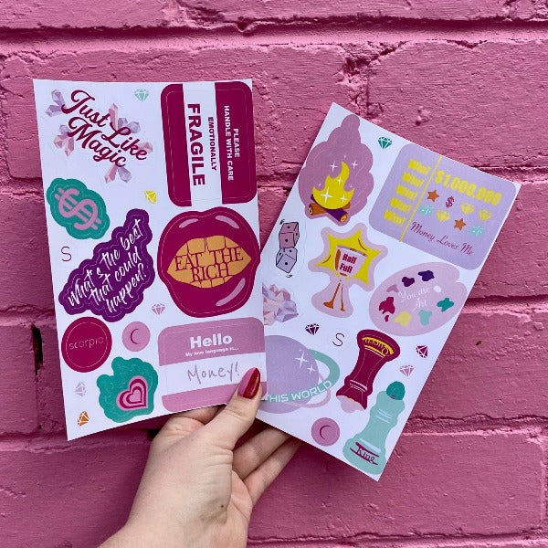 Two bold coloured fun sticker sheets held up in front of a pink brick wall. Featuring stickers such as Eat the Rich, Just like Magic, You are Art.