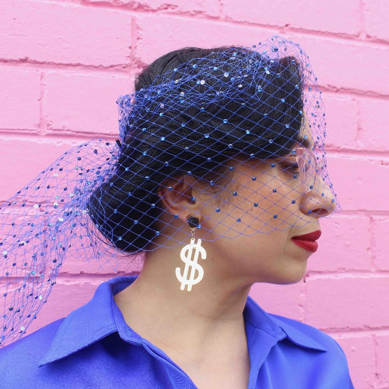 Woman in blue outfit stands in front of pink brick wall wearing "Dollar Dollar Bill" 18K Gold Statement Earrings shaped as a dollar sign featuring Black Obsidian rough cut crystals