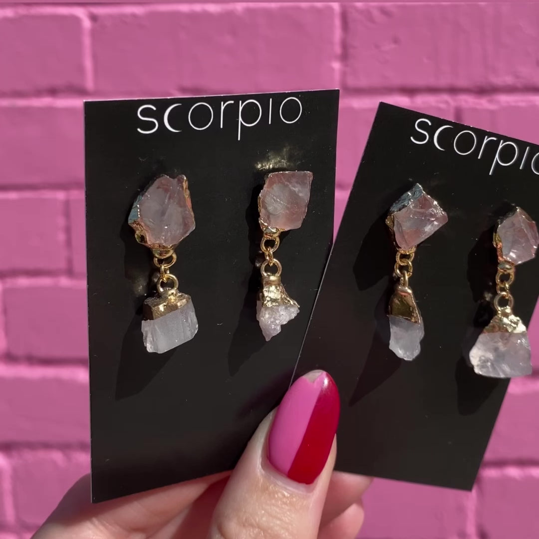 Two pairs of Crystal drop Earrings. 18K Gold Statement Earrings featuring Clear Quartz, Amethyst, Aquamarine, Black Obsidian, Lapis Lazuli rough cut crystals. On black Scorpio earring backing cards held in front of a pink brick wall.