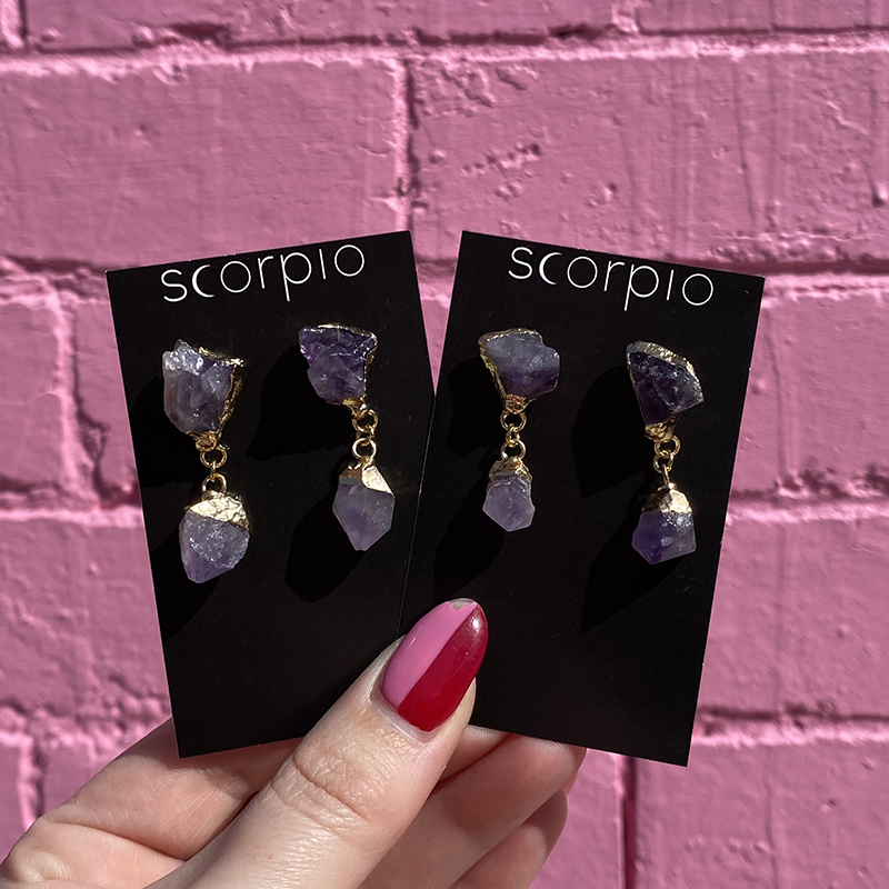 Two pairs of Crystal drop Earrings. 18K Gold Statement Earrings featuring Amethyst rough cut crystals. On black Scorpio earring backing cards held in front of a pink brick wall.