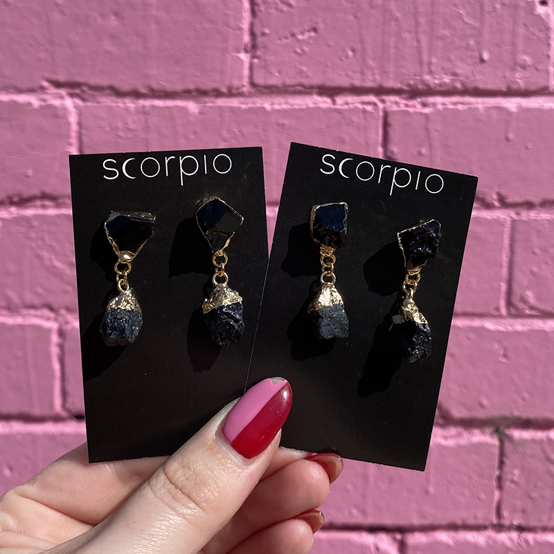 Two pairs of Crystal drop Earrings. 18K Gold Statement Earrings featuring Black Obsidian rough cut crystals. On black Scorpio earring backing cards held in front of a pink brick wall.