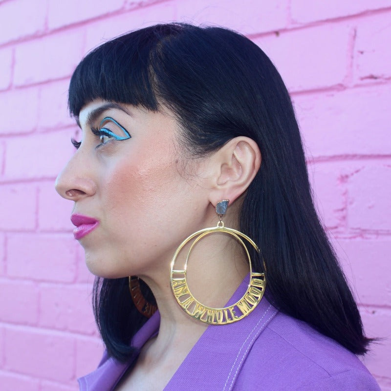 18K Gold Statement Earrings of a circular shaped design with the words "I am a whole mood!" wrapped around the edges. featuring Aquamarine rough cut crystals. Modelled by Margeaux in front of a pink brick wall.