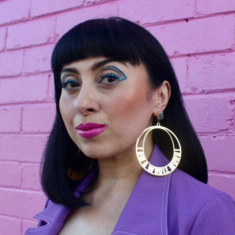 18K Gold Statement Earrings of a circular shaped design with the words "I am a whole mood!" wrapped around the edges. featuring Aquamarine rough cut crystals. Modelled by Margeaux in front of a pink brick wall.