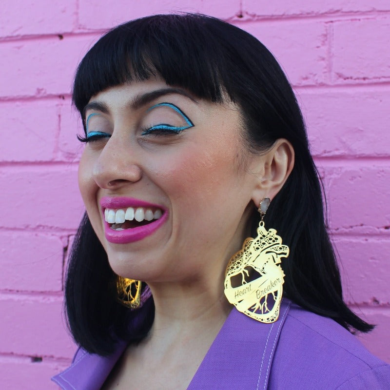 18K Gold Statement Earrings shaped as an anatomical heart with a banner wrapped around the middle reading "Heart Breaker" featuring Clear Quartz rough cut crystals. Modelled by Margeaux in front of a pink brick wall.