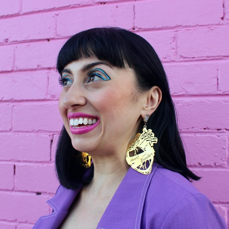 18K Gold Statement Earrings shaped as an anatomical heart with a banner wrapped around the middle reading "Heart Breaker" featuring Clear Quartz rough cut crystals. Modelled by Margeaux in front of a pink brick wall.