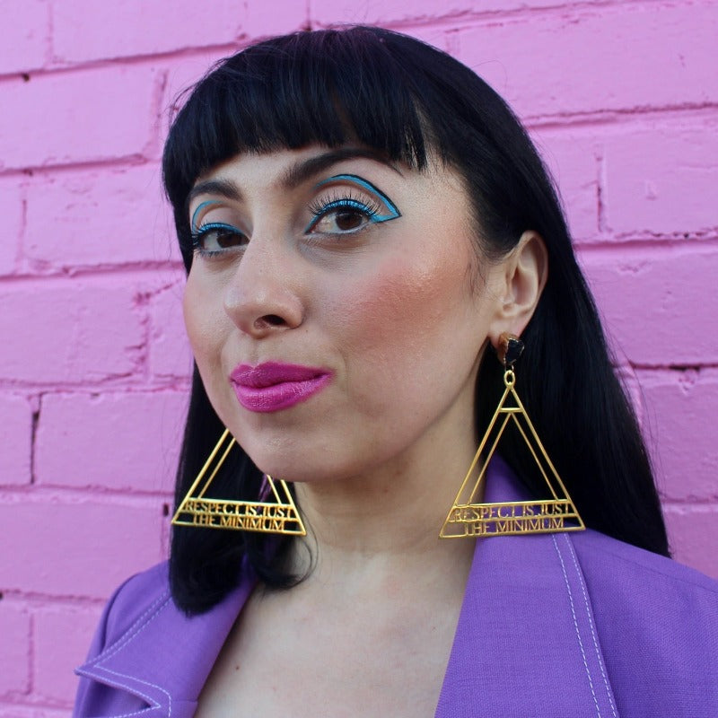 18K Gold Statement Earrings of a geometric triangular shaped design with the words "Respect is just the minimum" along the bottom edge of triangle. featuring Black Obsidian rough cut crystals. Modelled by Margeaux in front of a pink brick wall.