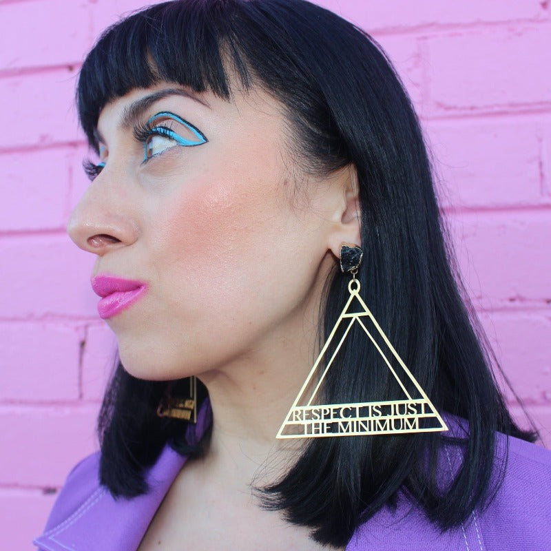 18K Gold Statement Earrings of a geometric triangular shaped design with the words "Respect is just the minimum" along the bottom edge of triangle. featuring Black Obsidian rough cut crystals. Modelled by Margeaux in front of a pink brick wall.