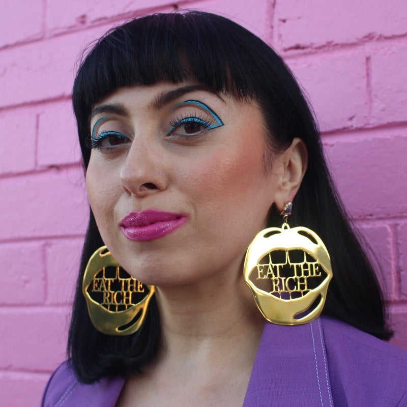 18K Gold Statement Earrings shaped as a mouth with the words "Eat The Rich" between the top and bottom row of teeth featuring Amethyst rough cut crystals. Modelled by Margeaux in front of a pink brick wall.