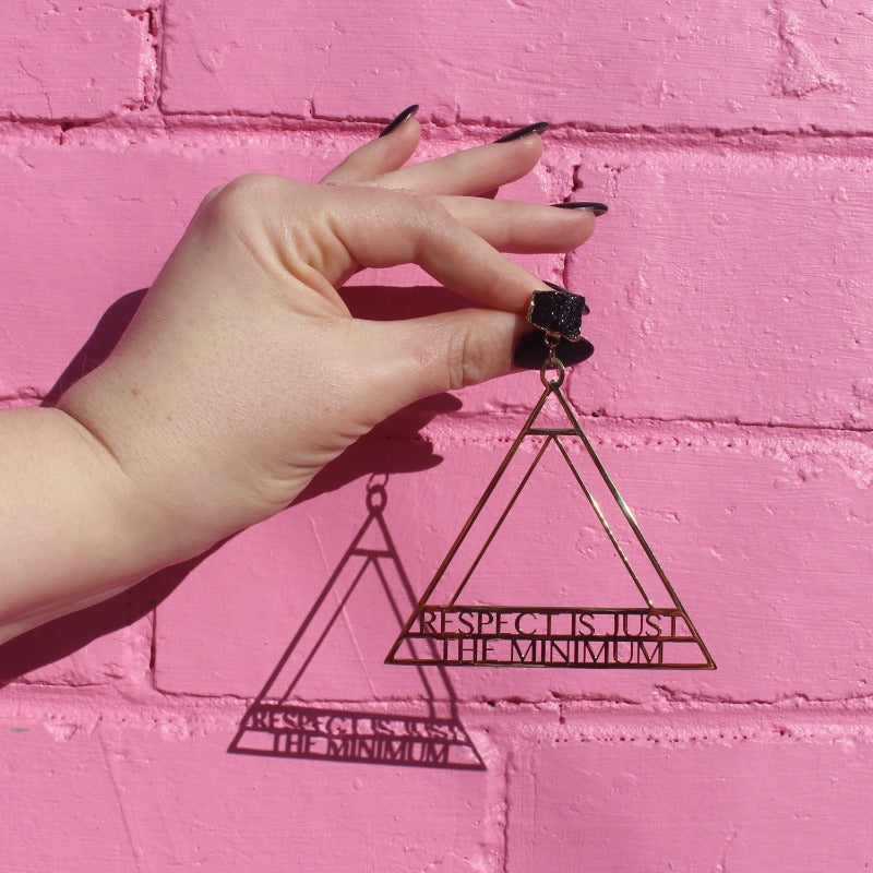 18K Gold Statement Earrings of a geometric triangular shaped design with the words "Respect is just the minimum" along the bottom edge of triangle. featuring Black Obsidian rough cut crystals. Held in front of a pink brick wall.