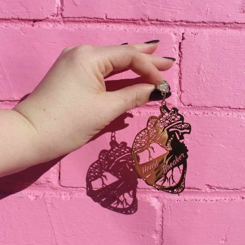 18K Gold Statement Earrings shaped as an anatomical heart with a banner wrapped around the middle reading "Heart Breaker" featuring Clear Quartz rough cut crystals. Held in front of a pink brick wall.