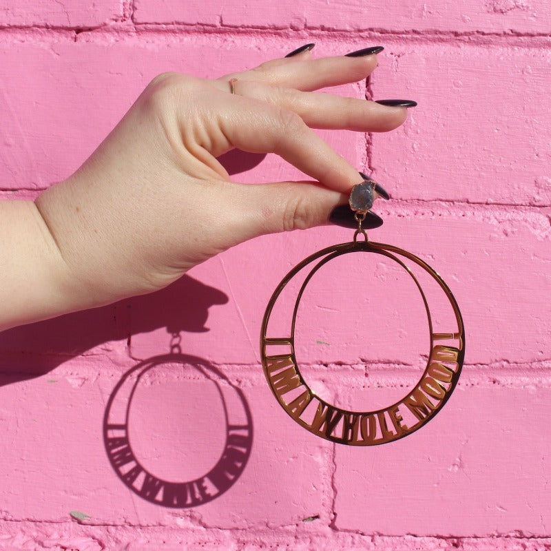 18K Gold Statement Earrings of a circular shaped design with the words "I am a whole mood!" wrapped around the edges. featuring Aquamarine rough cut crystals. Held in front of a pink brick wall.