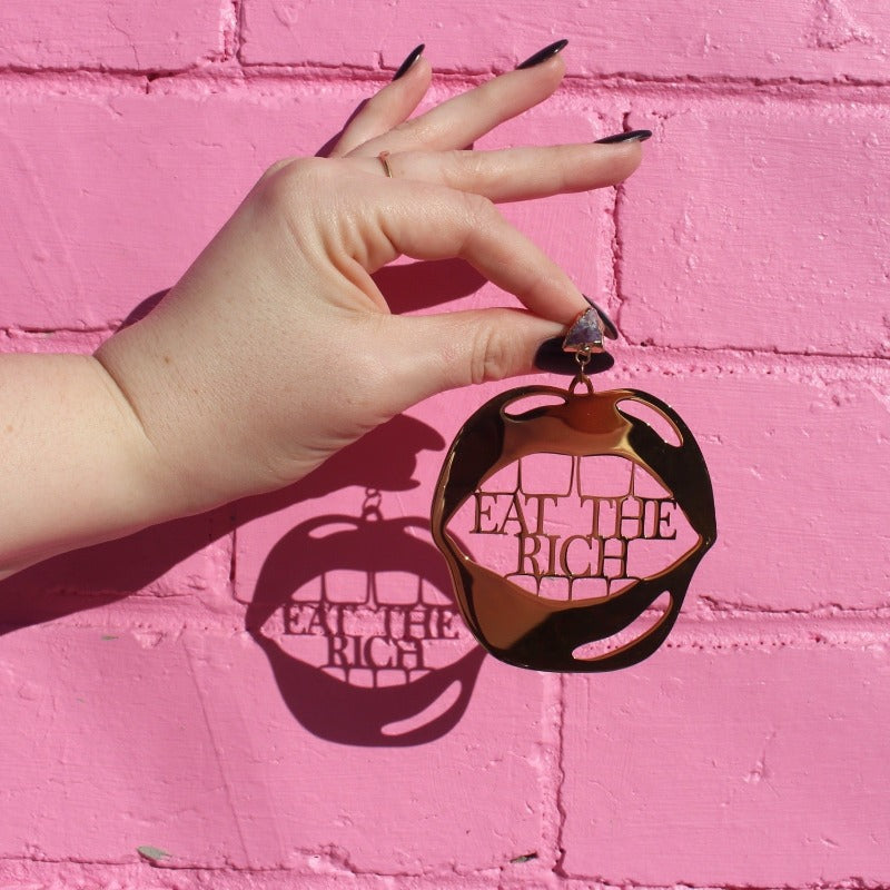 18K Gold Statement Earrings shaped as a mouth with the words "Eat The Rich" between the top and bottom row of teeth featuring Amethyst rough cut crystals. Held in front of a pink brick wall.