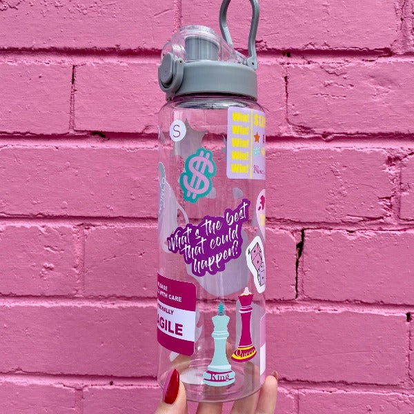 Two bold coloured fun sticker on a water bottle held up in front of a pink brick wall. Featuring stickers such as Eat the Rich, Just like Magic, You are Art.