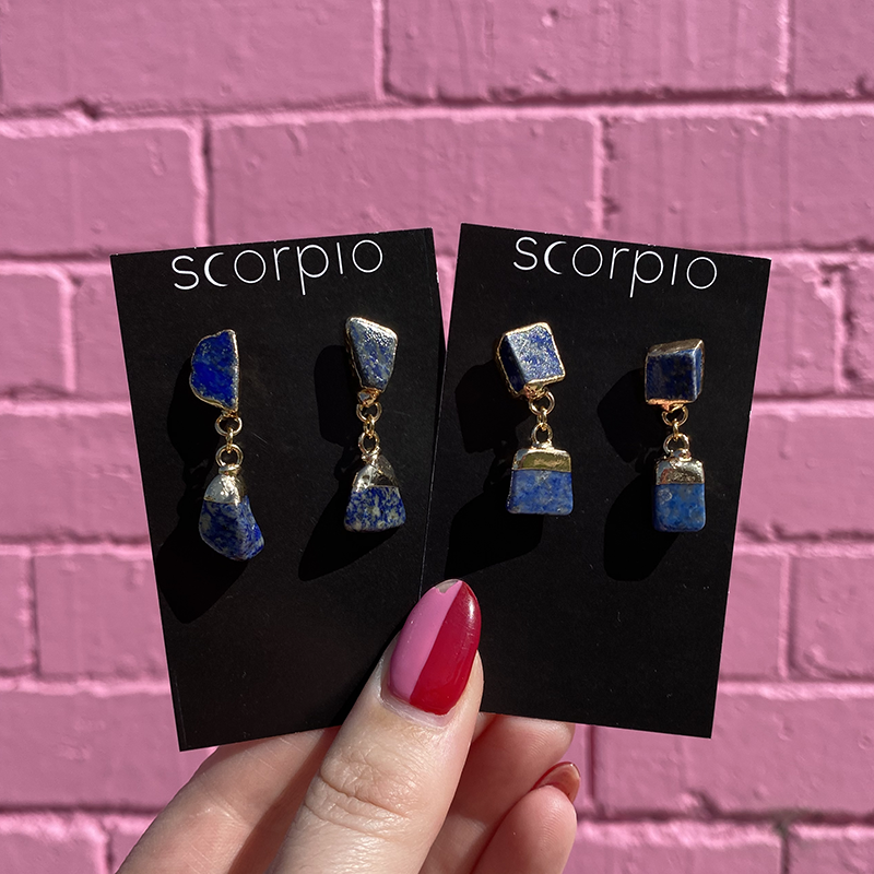 Two pairs of Crystal drop Earrings. 18K Gold Statement Earrings featuring Lapis Lazuli rough cut crystals. On black Scorpio earring backing cards held in front of a pink brick wall.